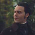 Ein Mann mit dunklem Haar blickt ernst zur Seite, vor einem grünen Hintergrund mit Bäumen.