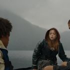 Drei Personen in einem kleinen Boot, tragen Regenkleidung. Sie wirken angespannt, Hintergrund zeigt neblige Berge und Wasser.