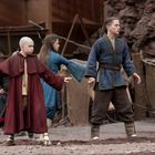 Personen mit Kampfszenenpose in traditionellen Gewändern, rustikale Umgebung mit Zelten und Felsen im Hintergrund.