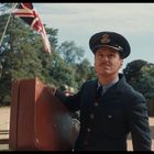 Ein Offizier in Uniform trägt einen Koffer. Eine britische Flagge weht im Hintergrund. Bäume umrahmen die Szene.