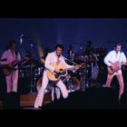 Eine Band in weißen Anzügen spielt auf einer Bühne, der Sänger in der Mitte hat eine Gitarre.