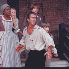 Eine Gruppe von Menschen in historischen Kleidern wirkt besorgt und ängstlich vor einem Backsteingebäude sowie einer Treppe.