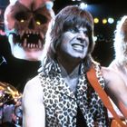 Ein Musiker mit langen Haaren und Leopardenweste spielt Gitarre. Im Hintergrund ein großes Totenkopf-Requisit.