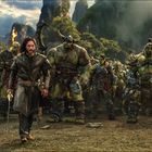 Ein Mann mit langem Haar steht vor einer Gruppe großer, muskulöser Fantasy-Kreaturen in rüstungsähnlicher Kleidung.