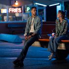 Neonschrift "FRIES" im Hintergrund. Zwei Personen sitzen auf der Ladefläche eines Trucks und unterhalten sich bei Nacht.