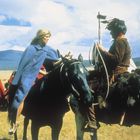 Zwei Personen sitzen auf Pferden und unterhalten sich. Im Hintergrund sind weitere Personen und Tipis auf einer Wiese.