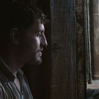 Keine Texte vorhanden. Ein Mann im Profil schaut aus einer dunklen Öffnung auf eine Bühne, auf der zwei Personen interagieren.