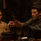 Zwei Personen in historischer Kleidung, eine Frau und ein Mann, betrachten ein altes Aufnahmegerät in einem dunklen Raum.