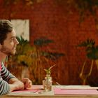 Zwei Männer sitzen an einem Tisch in einem gemütlichen Innenhof. Der Hintergrund ist mit Pflanzen und Ziegelwänden dekoriert.