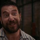 Ein Mann mit Bart und gestreiftem Hemd lächelt in einem dunklen, grob strukturierten Hintergrund.