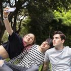 Drei junge Menschen sitzen auf Gras. Eine Person hat rosa Haare und macht ein Selfie. Die Atmosphäre ist fröhlich.