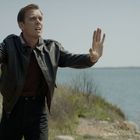 Ein Mann in Lederjacke hält eine Pistole, hebt die andere Hand, steht auf einer Wiese am Seeufer, besorgt.