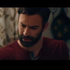 Keine sichtbaren Texte. Ein Mann mit Bart und rotem Shirt schaut nachdenklich, im Hintergrund Vorhang mit Blattmuster.