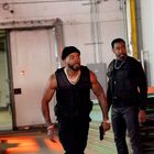 Zwei Männer in schwarzen taktischen Westen in einem Industrieraum, einer trägt eine Mütze und Ketten, beide wirken konzentriert.