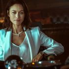 Eine Frau in einem eleganten weißen Blazer sitzt an einem Tisch, trägt eine Halskette und schaut selbstbewusst.