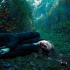 Keine sichtbaren Texte. Frau liegt in einem Waldweg, umgeben von Herbstlaub und Bergen im Hintergrund.