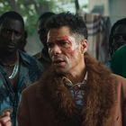 Eine Gruppe von Menschen, im Fokus ein Mann mit Kratzern im Gesicht, trägt einen Mantel mit Fellkragen, wirkt intensiv.