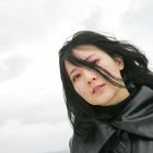 Keine Texte zu sehen. Frau mit langem, schwarzem Haar, schwarzer Lederjacke und rotem Lidschatten vor bewölktem Himmel.