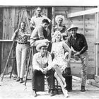 Sieben Personen in Cowboy-Kleidung stehen und sitzen vor einer Holzwand. Eine Frau im gepunkteten Kleid lächelt.