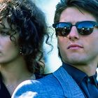 Eine Frau mit lockigem Haar steht neben einem Mann in Sonnenbrille und blauem Anzug. Beide blicken ernst zur Seite.