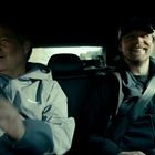 Zwei Männer sitzen lächelnd im Auto. Links trägt einer eine graue Jacke, daneben ein Mann mit Basecap. Sie wirken entspannt.