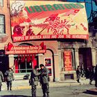 Großes Banner: "LIBERATORS. PEOPLE OF AMERICA ARISE AND JOIN THE REVOLUTION." Gebäude mit Soldaten davor.