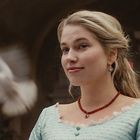 Ich kann dir nicht sagen, wer die Person ist. Eine Frau mit blondem Haar, roten Ohrringen und Halskette lächelt leicht. Eine Taube fliegt vorbei.