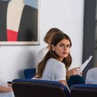 Keine erkennbaren Texte. Vier Frauen sitzen in einem Warteraum, eine schaut nachdenklich; an der Wand hängt ein abstraktes Gemälde.