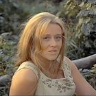 Keine sichtbaren Texte. Eine Frau mit hellem Haar schaut in die Kamera, im Hintergrund sind unscharfe Blätter.