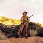 Es gibt keinen erkennbaren Text. Ein Cowboy steht in einer Wüstenlandschaft, hält ein Gewehr und trägt Hut und Mantel.