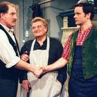 Zwei Männer schütteln sich die Hand, ein dritter schaut lächelnd zu. Alle tragen traditionell wirkende Kleidung und Schürzen.