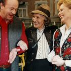 Zwei Frauen und ein Mann lächeln einander an. Die Frau in der Mitte trägt einen Hut. Alle sind in traditioneller Kleidung.