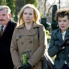 Drei Personen in eleganter Kleidung stehen draußen auf einem Friedhof. Eine Frau hält Blumen in der Hand.