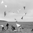 Fünf Personen rennen über ein Feld und werfen Drachen in die Luft. Der Himmel ist bewölkt, und die Szenerie wirkt dynamisch.