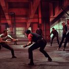 Keine Texte sichtbar. Zwei Personen in einem kämpferischen Duell, umgeben von einer Gruppe, in einem industriellen Raum.