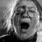 Eine ältere Person mit runder Brille schreit mit offenem Mund. Auf dem Gesicht sind blutähnliche Spuren zu sehen.