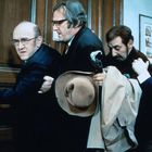 Vier Männer drängen sich an einer Tür; einer hält einen Hut und Mantel, alle wirken angespannt.