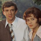 Ein Mann im Anzug und eine Frau mit gepunkteter Bluse, beide draußen in einem grünen Park.