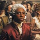 Ein Mann in Rokokokleidung mit gepuderter Perücke sitzt inmitten einer festlich gekleideten Menge.