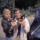 Zwei Männer richten Pistolen nach vorne, stehen vor einem schwarzen Auto, im Hintergrund Bäume und Büsche.