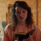 Ein Mädchen mit braunem lockigem Haar liest ein Buch in einem Zimmer mit orange-blumiger Tapete.