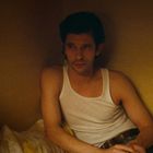 Ich sehe keinen Text. Ein Mann in einem weißen Unterhemd sitzt nachdenklich auf einem Bett in einem schwach beleuchteten Raum.