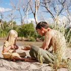 Ein Mädchen und ein Mann sitzen am sandigen Flussufer, umgeben von Bäumen. Das Mädchen trägt braune Fäustlinge.