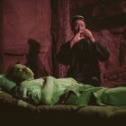 Kein Text erkennbar. Zwei Personen, eine liegt, die andere spielt eine Flöte. Dunkle, düstere Umgebung, mystische Atmosphäre.