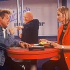 Zwei Männer und eine Frau an einem runden Tisch. Ein Mann zeigt, die Frau trägt einen roten Blazer und hält Essen.