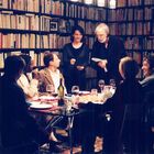 In einer Bibliothek sitzen sechs Menschen an einem Tisch mit Wein und Speisen, Bücherregale im Hintergrund.