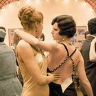 Paartanz in einem festlich beleuchteten Saal. Frauen in eleganten Kleidern tanzen eng umschlungen, umgeben von weiteren Paaren.