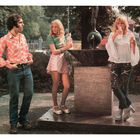 Drei Personen stehen an einem Brunnen. Zwei Frauen mit Getränken, eine in einer rosa Bluse, die andere im Rock. Ein Mann in Hemd.