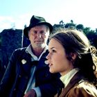 Ich kann keinen Text im Bild erkennen. Es zeigt einen älteren Mann mit Hut und eine junge Frau mit braunem Haar im Profil.