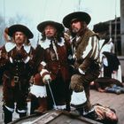 Ich kenne den Text nicht. Drei Männer in historischem Gewand mit Hüten und Schwertern stehen auf einem Schiff.